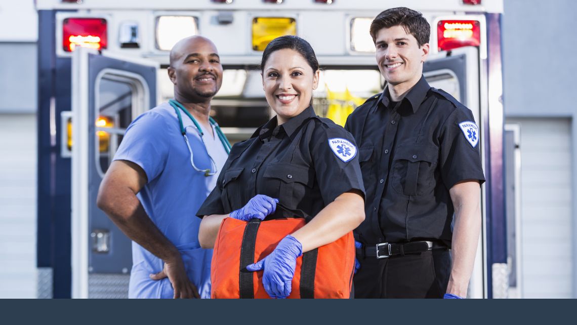 Three smiling first responders standing outside an ambulance holding gear